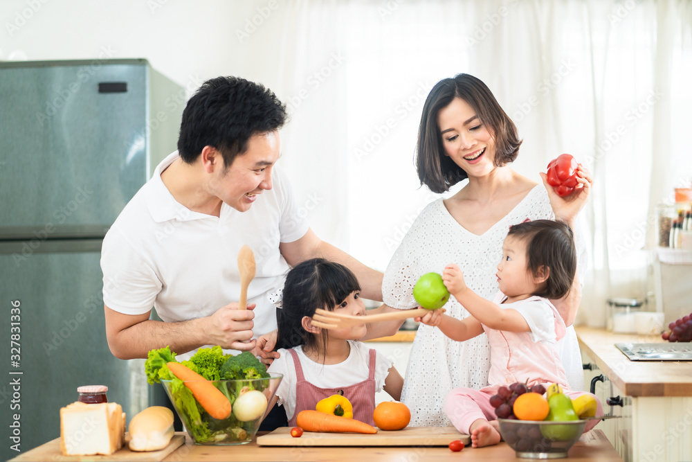 Lovely cute Asian family making food in kitchen at home. Portrait of smiling mother, dad and children standing at cooking counter. kid feeding dad some fruit with smile. Happy family activity together