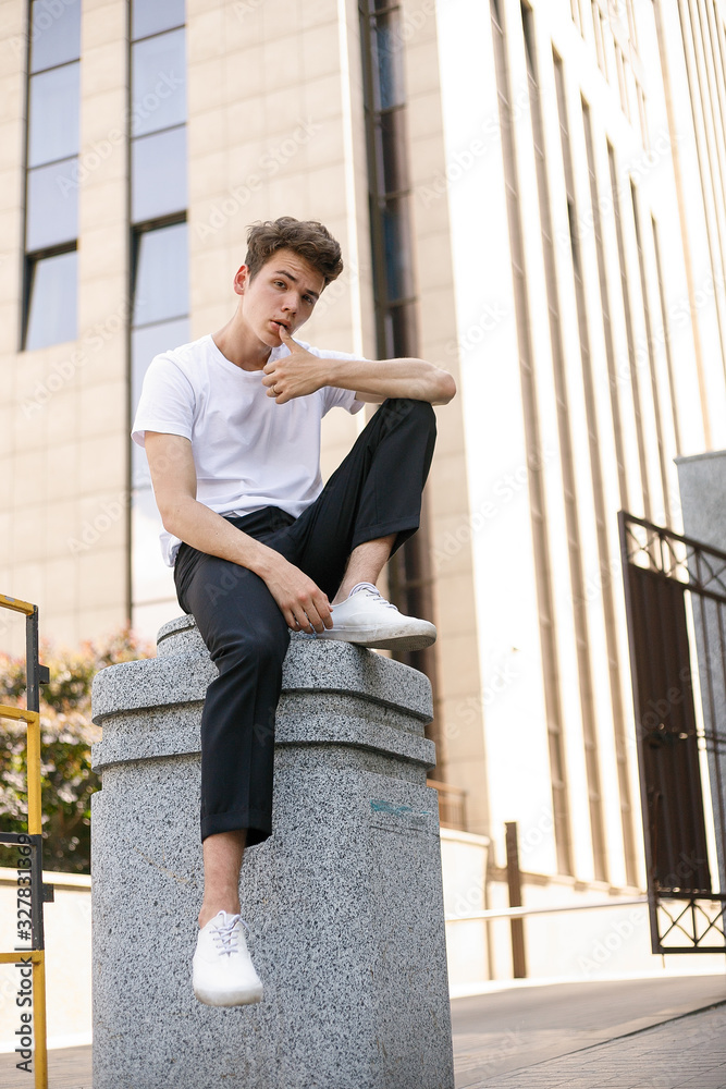 Elegant young man in a fashionable black shirt in a white stylish t-shirt in black pants with a trendy hairstyle rests near a modern business center. Attractive guy in the street on a summer day.