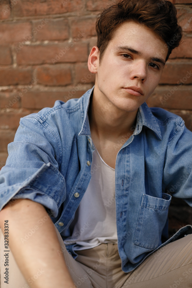 Young hipster sits in his blue denim button up shirt showing an unlabeled white cotton t-shirt against a brick wall background