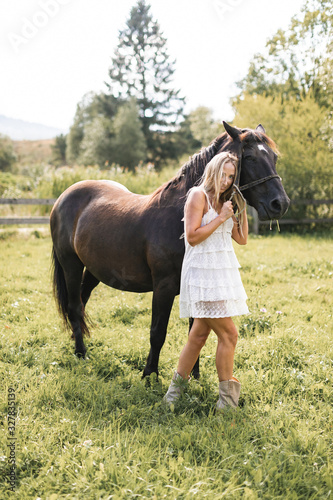 Horse and girl outdoors. Lovely stylish cowgirl with her horse on a ranch. Beautiful girl in white short boho dress with black horse in summer field at sunset.