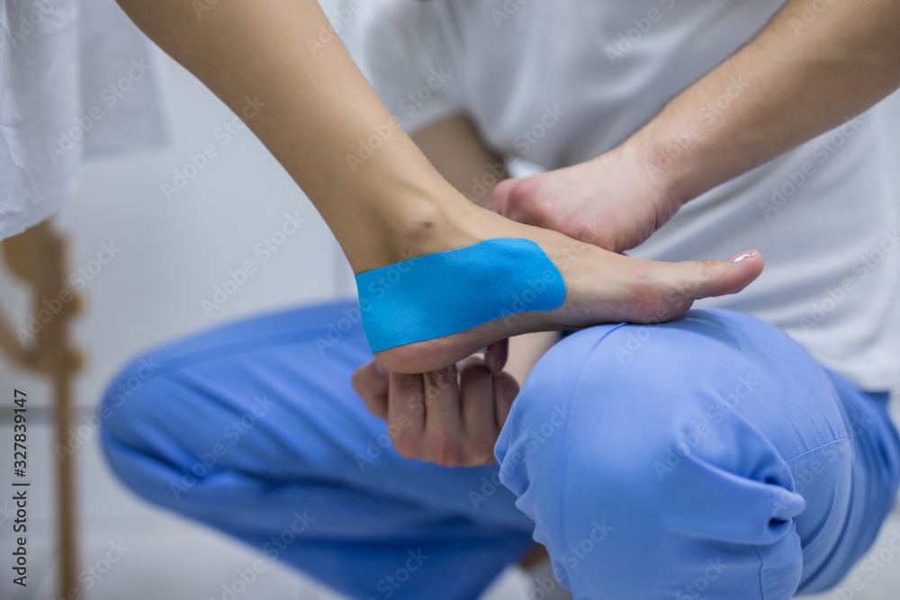 Young man physiotherapist is applying kinesio tape to a patient's leg
