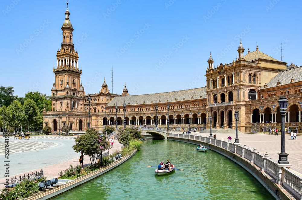 Seville, Spain - Square of Spain, a neo-Moorish-style urban ensemble, light brown buildings with numerous arches, people in boats sailing along a channel with greenish water, in the summer afternoon.