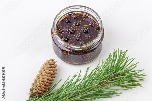 Unusual jam from pine cones in glass jar on white background among pine branches and cones. Organic and vegetarian sweet dessert close up  copy space for text. Remedy for colds