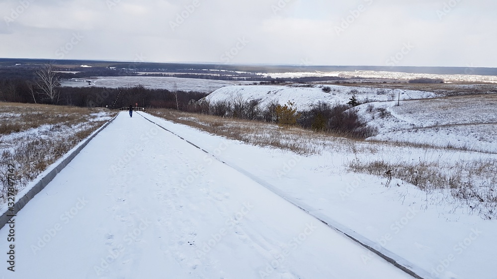Snowy winter road among hills, fields and a small forest on a cloudy February day Snowy winter road among hills, fields and a small forest on a cloudy February day