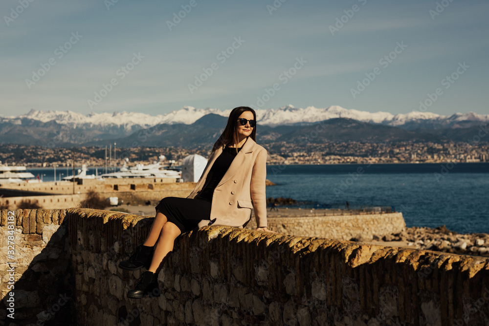 Young woman on the embankment of Antibes at french riviera. Lovely elegant girl in beige coat and black dress with glasses visiting the famous Antibes port. Young woman on the embankment of Antibes at french riviera. Lovely elegant girl in beige coat and black dress with glasses visiting the famous Antibes port.
