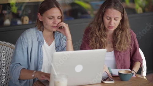 Happy women sitting at table in coffee shop and work together using laptop