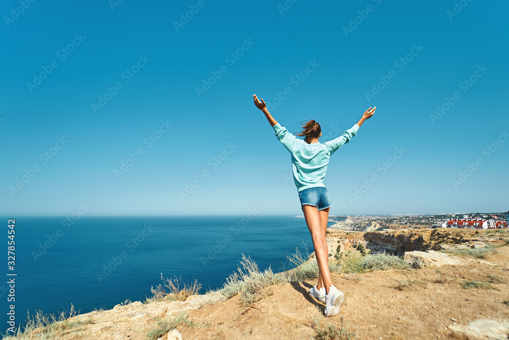 back view of carefree young woman standing with raised hands on cliff edge with beautiful sea view and enjoying wonderful nature. Full length portrait back view of carefree young woman standing with raised hands on cliff edge with beautiful sea view and enjoying wonderful nature. Full length portrait