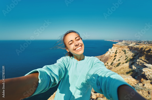 young female traveler making selfie on cliff edge with beautiful sea view, laughing and smiling to the camera, making funny face.