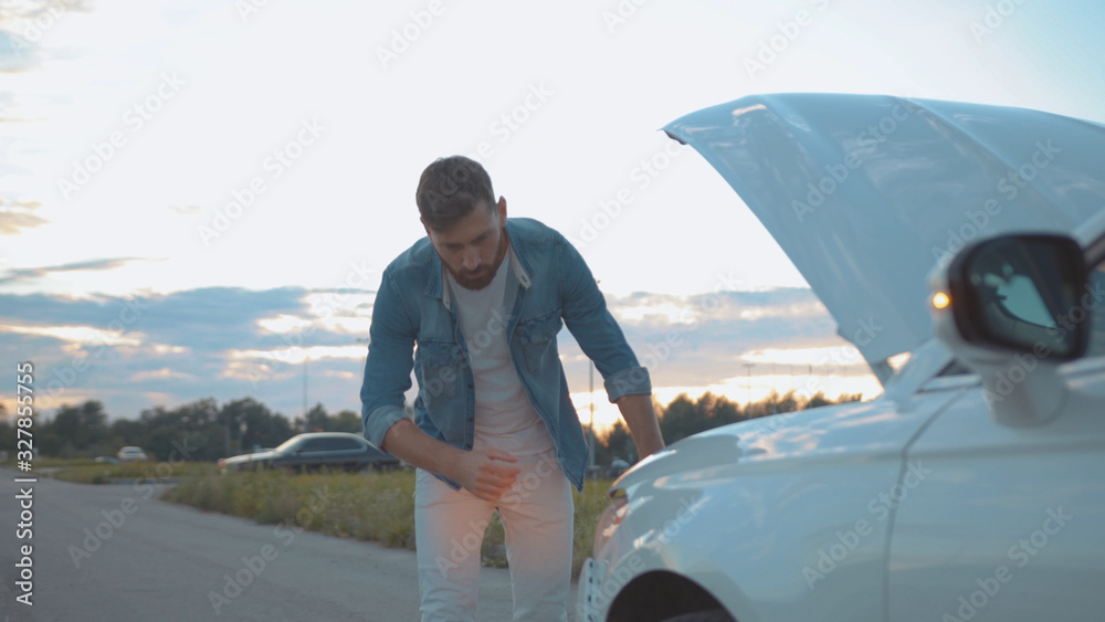 Portrait of disappointed male driver leaning on crashed car at roadside. Close-up confused young man in doubt having problems with a car on country road.