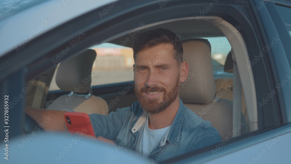 Handsome portrait of male driver holding mobile phone and steering wheel using device while sitting in modern auto on sunny street. Handsome portrait of male driver holding mobile phone and steering wheel using device while sitting in modern auto on sunny street.