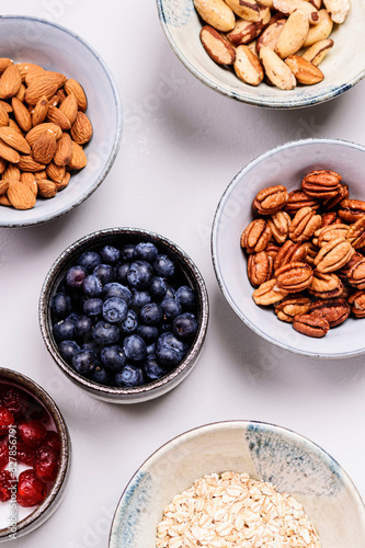 Ingredients for homemade granola: oatmeal, dried cherry, pecans, almond, brazilan nuts, blueberries in a bowls on grey concrete background. Healthy diet breakfast. Flatlay. Top view. Selective focus photo