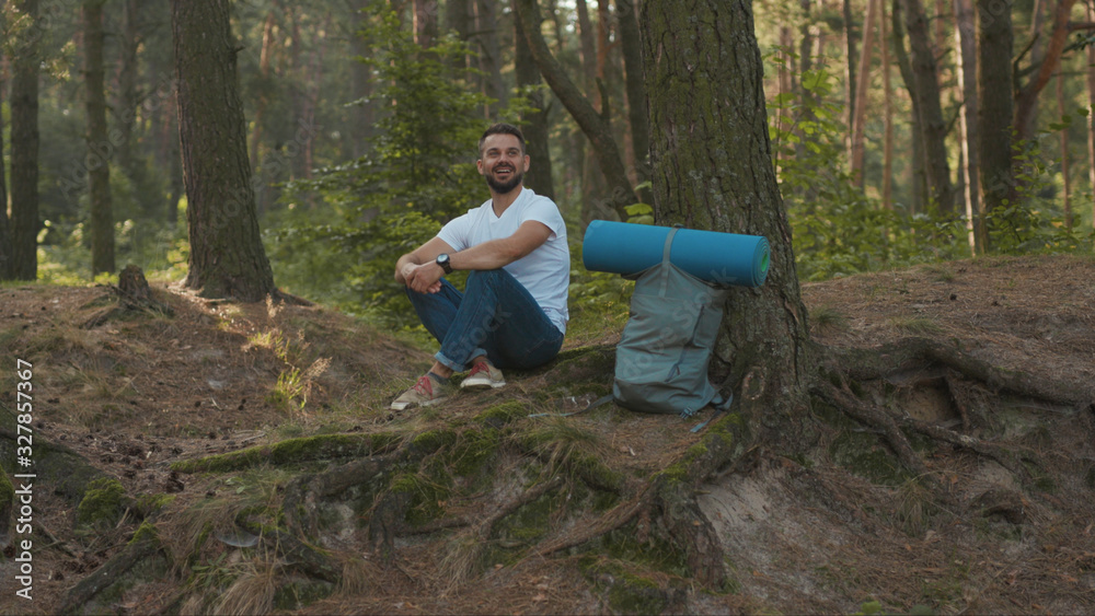 In the forest young man hiker sitting near tree look around smiling at sunlight nature alone hiking woods activity young vacation attractive autumn hiker travel slow motion In the forest young man hiker sitting near tree look around smiling at sunlight nature alone hiking woods activity young vacation attractive autumn hiker travel slow motion