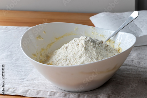A white bowl with cottage cheese dough, on which a hill of flour is poured, stands on a linen tablecloth on a wooden table. The process of making curd bagels or croissants