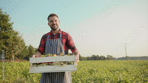 Close up handsome farmer is holding a box of organic vegetables walk look at camera at sunlight agriculture farm field harvest garden nutrition organic fresh outdoor slow motion