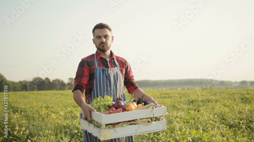 Close up handsome farmer is holding a box of organic vegetables walk look at camera at sunlight agriculture farm field harvest garden nutrition organic fresh outdoor slow motion