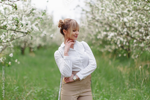 Beautiful young blonde woman in white shirt with backpack posing under apple tree in blossom in Spring garden