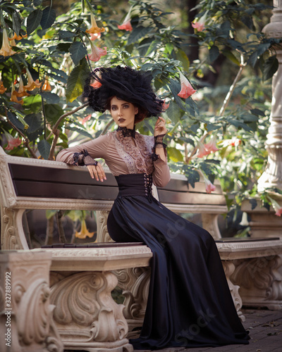 Portrait of amazing cute young woman in big lacy hat and vintage brown blouse with black long skirt sits on old stone carved bench in green park against background of large white and pink flowers