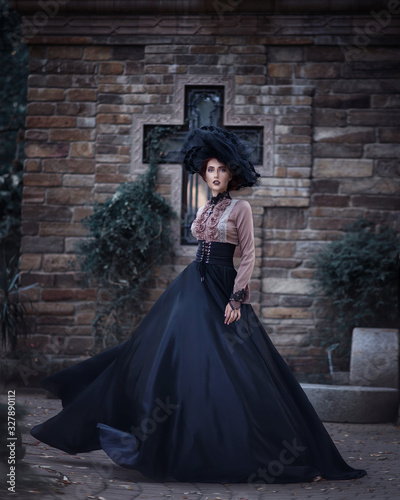 Portrait of amazing cute young woman in large lacy black hat and vintage brown blouse and black long flowing skirt stands and looks at camera on background dark brick wall