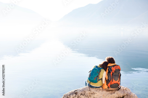 Couple travelers with hiking backpacks at sea beach. Tourists by blue lake and mountains. Young man and woman enjoying traveling, adventure, relaxation, silence and calmness. Copy space. Rear view. © Marina April