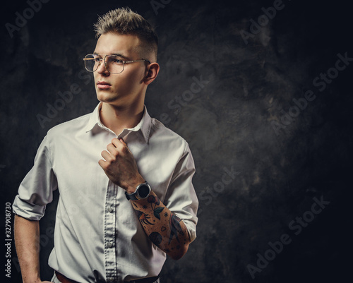 Young handsome student guy in a white shirt and glasses, with tattoos on his hand, wearing his watches in a dark studio isolated on grey background, looking cool and thoughtful