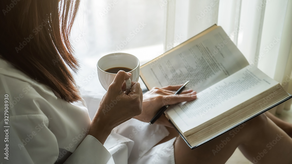 Obraz premium Asian woman holding cup of coffee and reading a book beside the window after get up in morning. Morning lifestyle concept.