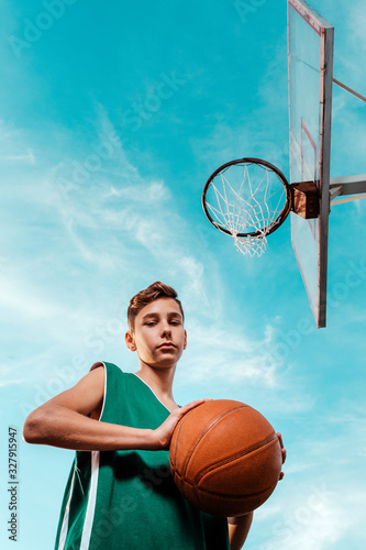 A young teenager in green sportswear poses with a basketball. Bottom view. The sky in the background. Copy space. Concept of sport and activity © _KUBE_