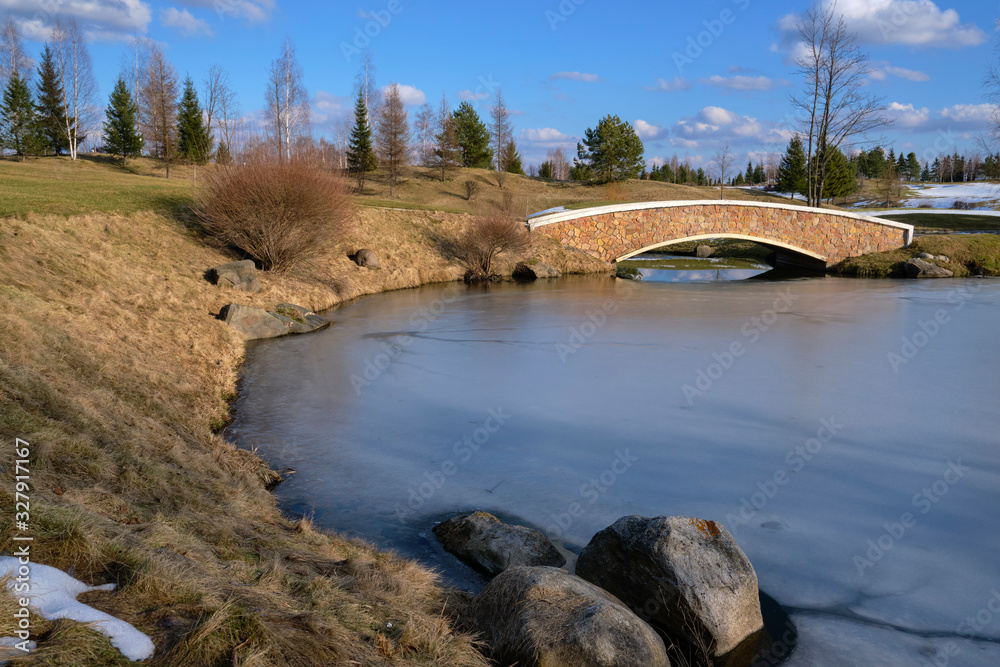  The landscape is early spring. The landscape of the city park with melting snow on hills, trees, shrubs, a bridge over the lake covered with ice and a blue sky with clouds.