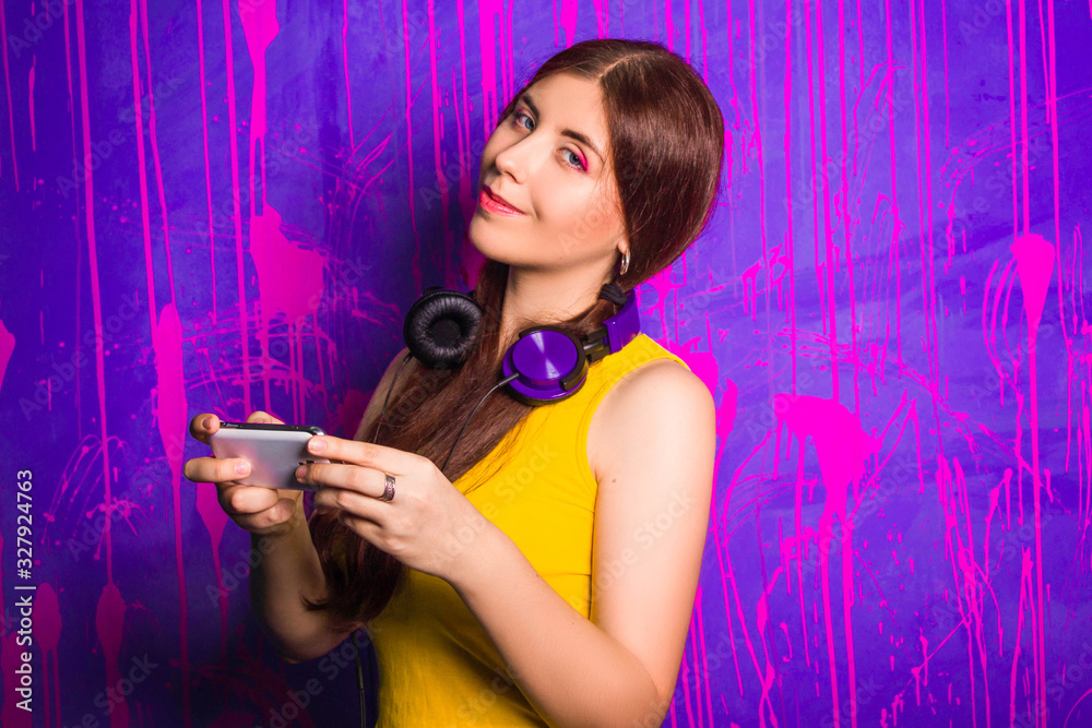 Attractive, beautiful woman with long hair and headphones on her neck, makes selfie while standing on an abstract, purple background. Attractive, beautiful woman with long hair and headphones on her neck, makes selfie while standing on an abstract, purple background.