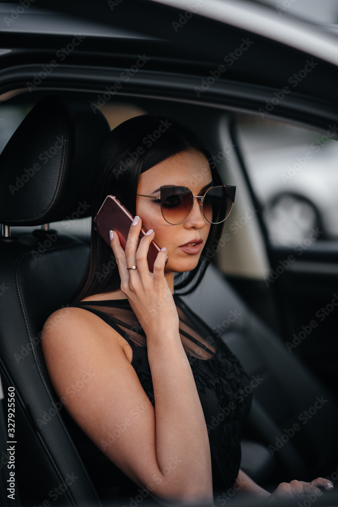 Stylish young girl sitting in a business class car in a black dress and talking on the phone. Business fashion and style
