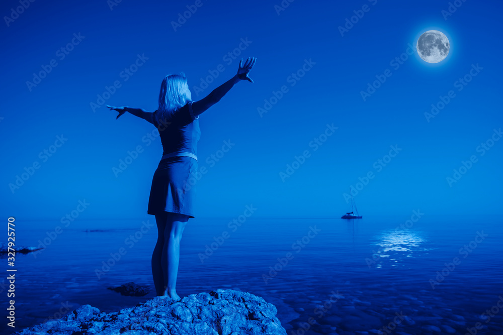 Rear view unidentified young happy woman stands on a stone raise Hands up Looking at the big moon and calm clear sea water against the backdrop of landscape and clear night sky. Full moon concept