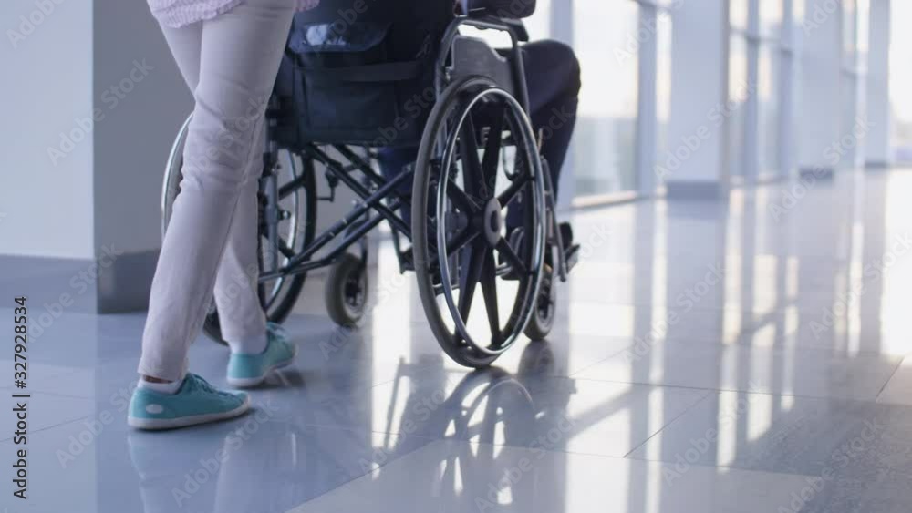 Low-section shotof nurse pushing driving wheelchair with handicapped patient through the corridor with mirror floor