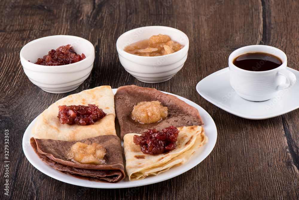 Pancakes on a white plate, apple jam of different varieties and coffee over a wooden background.