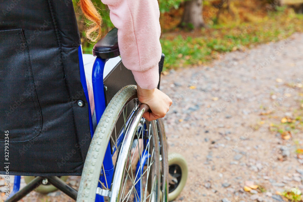 Naklejka premium Hand handicap woman in wheelchair wheel on road in hospital park waiting for patient services. Unrecognizable paralyzed girl in invalid chair for disabled people outdoors. Rehabilitation concept.