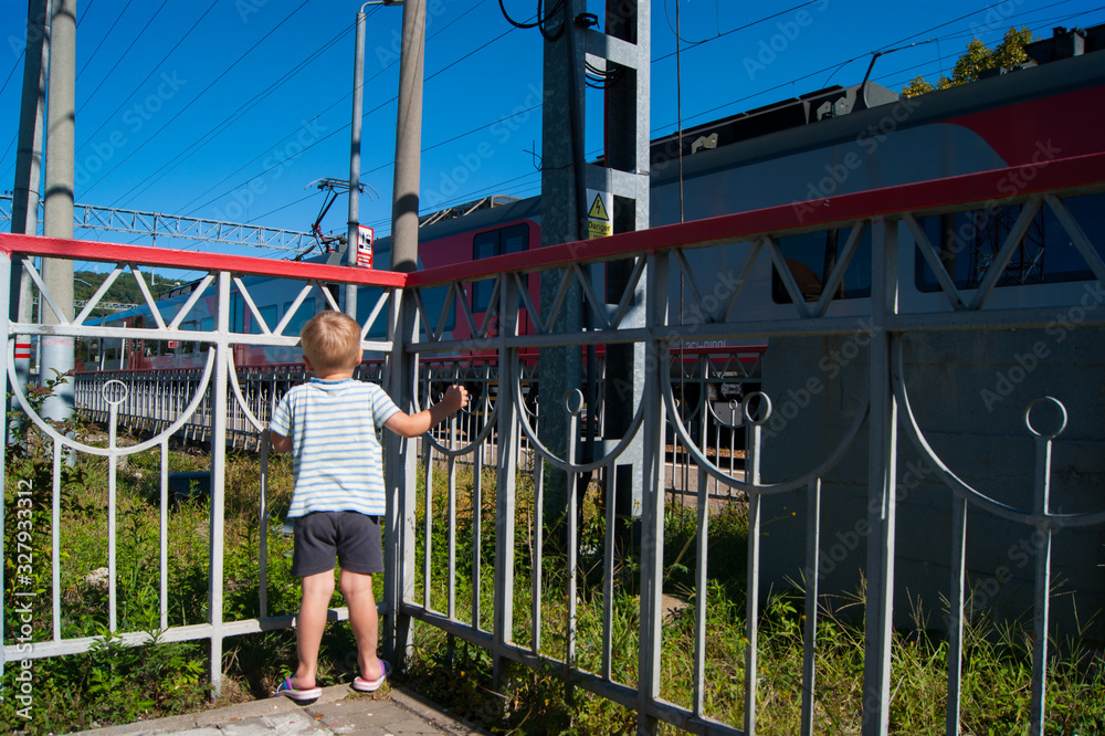 A little blond-haired boy of three years old stands near a fence near the railway and looks at a passing train.
