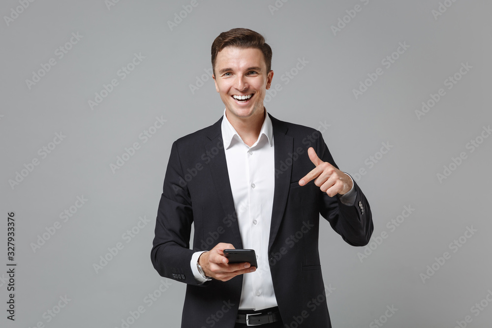 Funny young business man in classic black suit shirt posing isolated on grey wall background. Achievement career wealth business concept. Mock up copy space. Pointing index finger on mobile phone. Funny young business man in classic black suit shirt posing isolated on grey wall background. Achievement career wealth business concept. Mock up copy space. Pointing index finger on mobile phone.