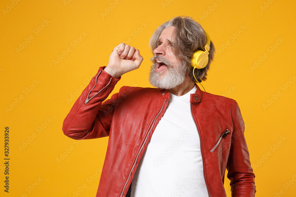 Elderly gray-haired mustache bearded man in red leather jacket posing isolated on yellow orange wall background. People lifestyle concept. Mock up copy space. Listen music in headphones, sing song. Elderly gray-haired mustache bearded man in red leather jacket posing isolated on yellow orange wall background. People lifestyle concept. Mock up copy space. Listen music in headphones, sing song.