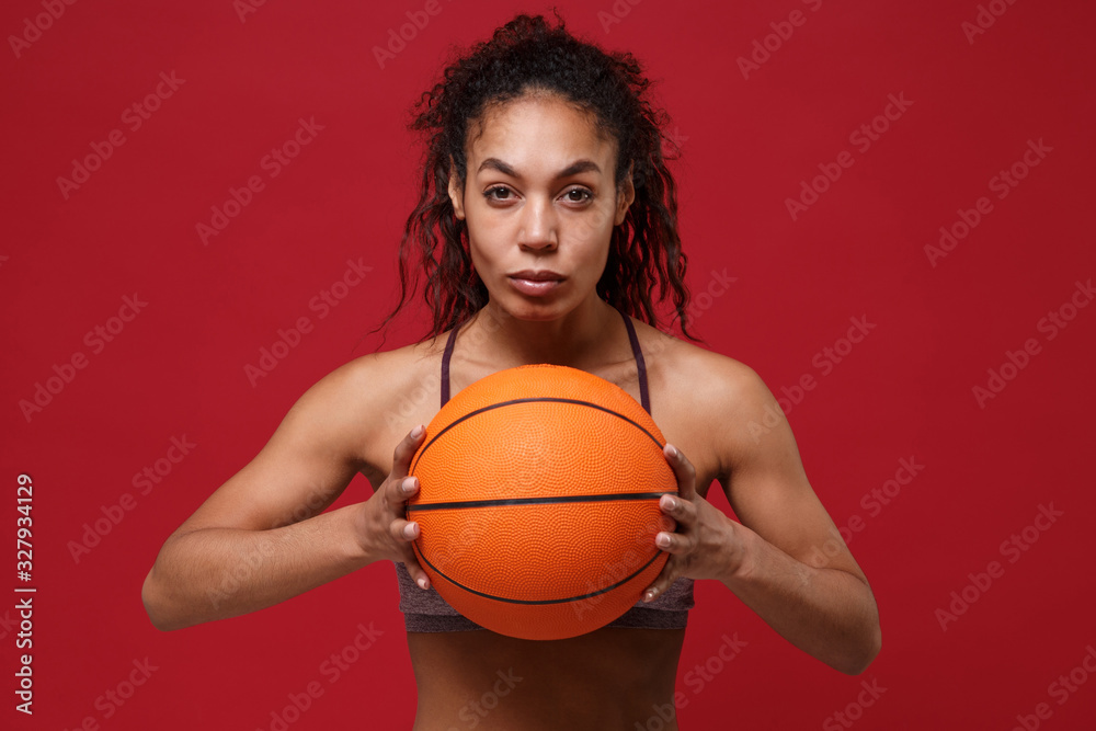 Confident young african american sports fitness basketball player woman in sportswear working out isolated on red background studio portrait. Sport exercises healthy lifestyle concept. Holding ball.