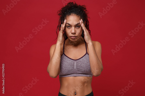 Tired sick young african american sports fitness woman in sportswear posing working out isolated on red wall background. Sport exercises healthy lifestyle concept. Put hands on head, having headache.