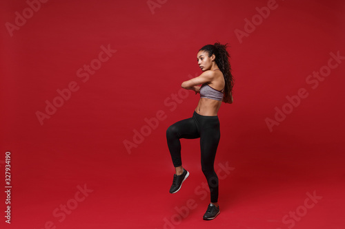 Beautiful young african american sports fitness woman in sportswear posing working out isolated on red background. Sport exercises healthy lifestyle concept. Doing stretching exercising looking aside. © ViDi Studio