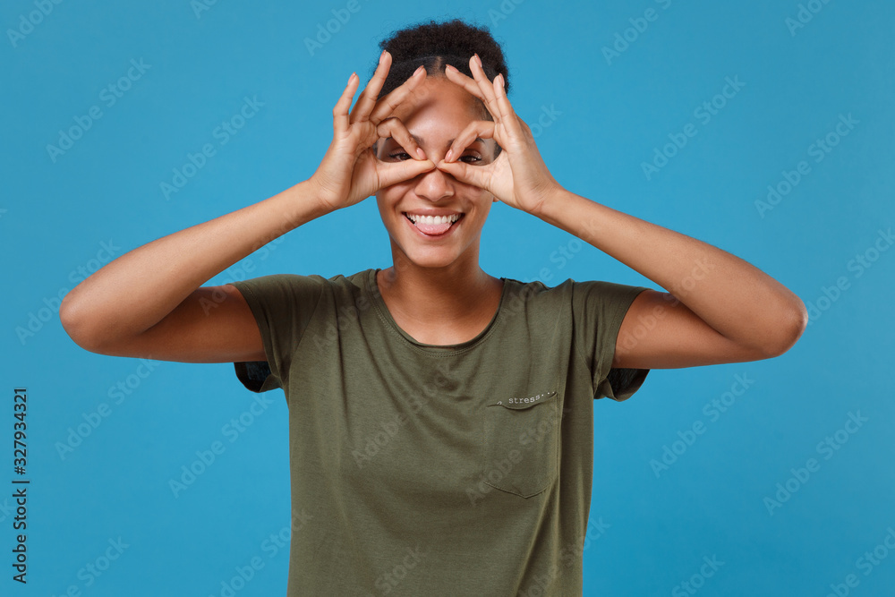 Funny young african american woman girl in casual t-shirt posing isolated on blue background. People lifestyle concept. Mock up copy space. Holding hands near eyes imitating eyeglasses or binoculars. Funny young african american woman girl in casual t-shirt posing isolated on blue background. People lifestyle concept. Mock up copy space. Holding hands near eyes imitating eyeglasses or binoculars.