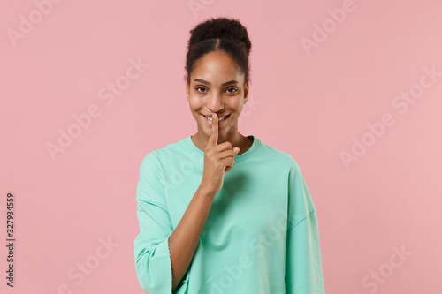 Smiling young african american girl in green sweatshirt posing isolated on pastel pink background. People lifestyle concept. Mock up copy space. Saying hush be quiet with finger on lips shhh gesture.