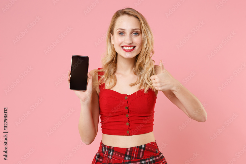 Naklejka premium Smiling young blonde woman girl in red sexy clothes posing isolated on pastel pink background. People lifestyle concept. Mock up copy space. Hold mobile phone with blank empty screen showing thumb up.
