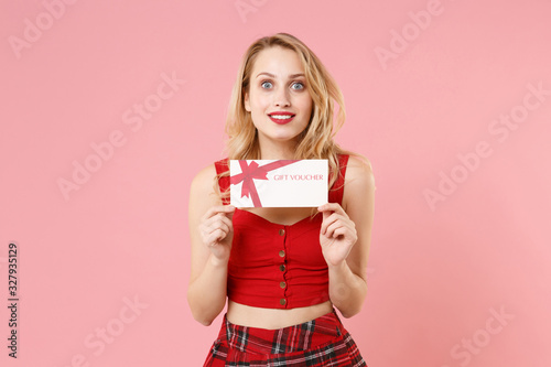 Excited young blonde woman girl in red sexy clothes isolated on pastel pink wall background studio portrait. People sincere emotions lifestyle concept. Mock up copy space. Holding gift certificate.