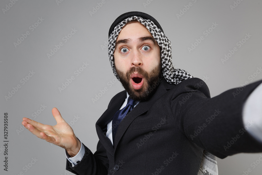 Fototapeta premium Close up of arabian muslim businessman in keffiyeh kafiya ring igal agal suit isolated on gray background. Achievement career wealth business. Doing selfie shot on mobile phone keeping mouth open.