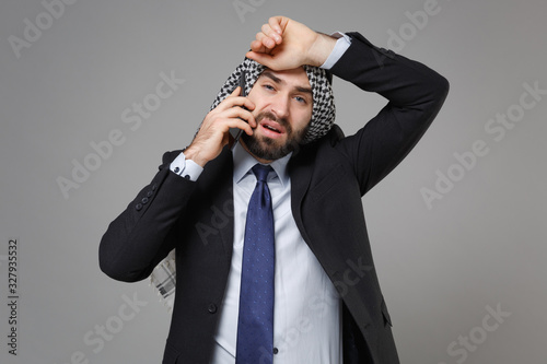 Tired bearded arabian muslim businessman in keffiyeh kafiya ring igal agal classic black suit shirt isolated on gray background. Achievement career wealth business concept. Talking on mobile phone. photo