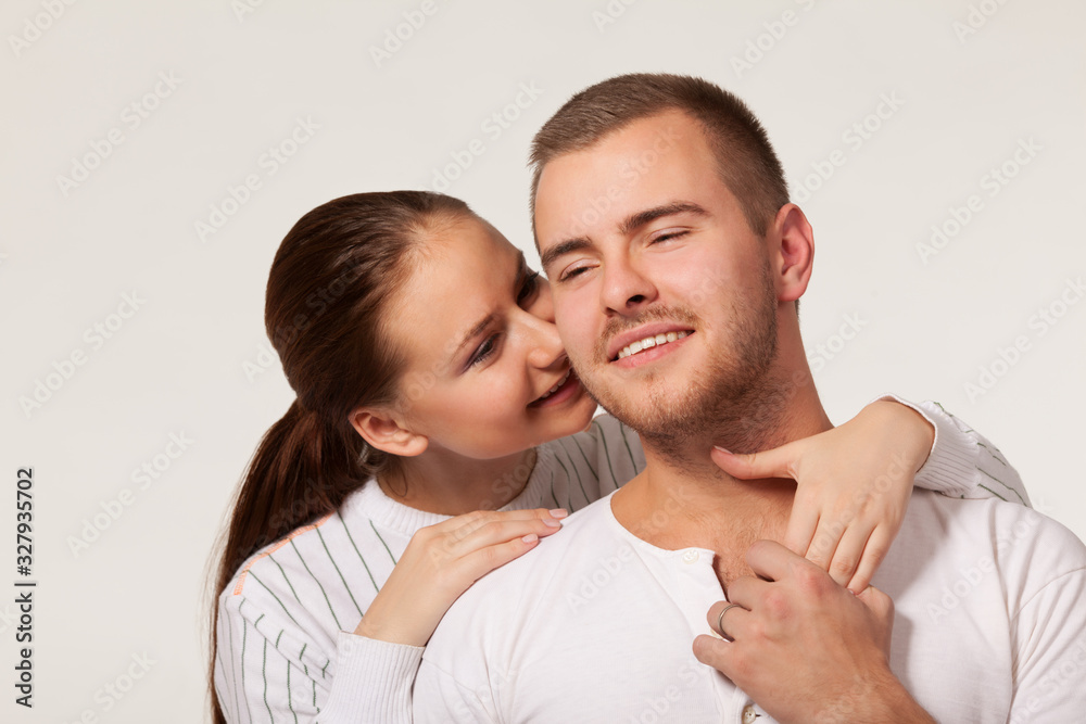 Front view of charming young woman hugging joyful guy from behind and biting ear. Handsome man holding girlfriend hand and smiling. Isolated on white studio background. Concept of relationships. Front view of charming young woman hugging joyful guy from behind and biting ear. Handsome man holding girlfriend hand and smiling. Isolated on white studio background. Concept of relationships.