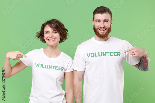 Smiling two young friends couple in white volunteer t-shirt isolated on pastel green background. Voluntary free work assistance help charity grace teamwork concept. Point index fingers on themselves.