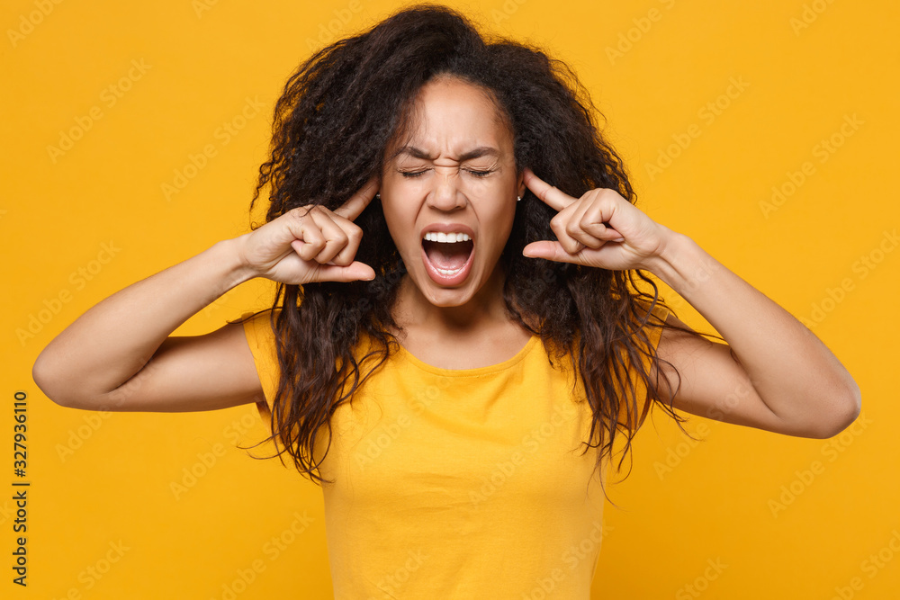 Crazy young african american woman girl in casual t-shirt posing isolated on yellow orange background. People lifestyle concept. Mock up copy space. Covering ears with fingers, keeping eyes closed. Crazy young african american woman girl in casual t-shirt posing isolated on yellow orange background. People lifestyle concept. Mock up copy space. Covering ears with fingers, keeping eyes closed.