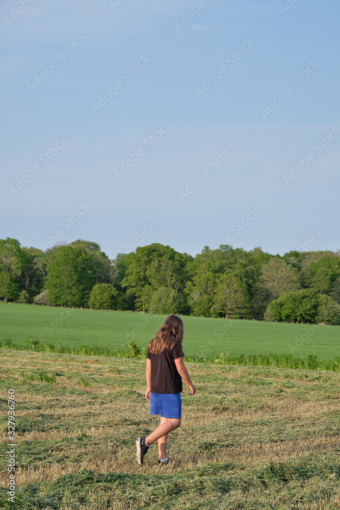 Boy with long hair, black t-shirt and blue shorts walking on a green field with trees in the background.