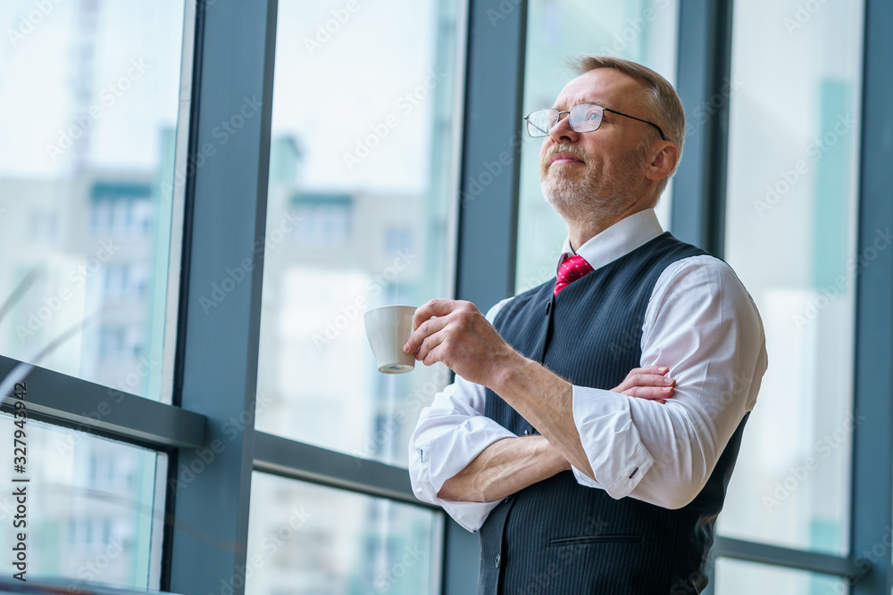 Taking time for coffee break. Confident mature man in formalwear holding coffee cup and looking away while standing near the window Taking time for coffee break. Confident mature man in formalwear holding coffee cup and looking away while standing near the window